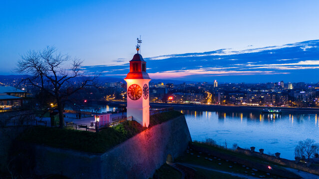 Aerial view of a clock tower bathed in a warm red glow stands prominently against the cool blue twilight sky overlooking the Danube River, Petrovaradin, Vojvodina, Serbia.