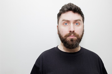 Fototapeta premium A direct, neutral headshot portrait of a man with dark hair, beard and blue eyes in a black t-shirt on a white background. Stock photo