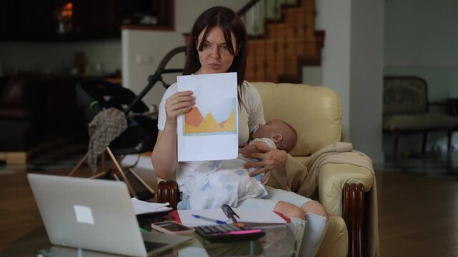 Busy mother holding her sleeping newborn baby while participating in an online video call, presenting a chart and working on her laptop