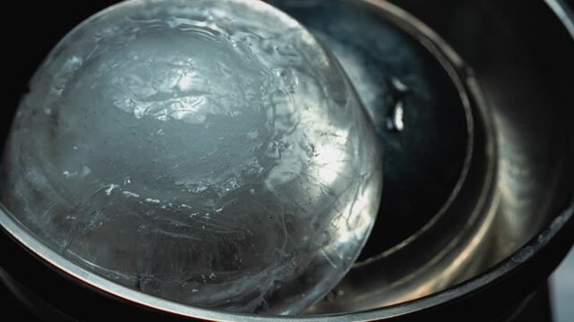 Timelapse crystal-clear ice sphere melting. A large piece of ice, a hemisphere, melts in a metal plate. Mesmerizing macro view of ice transformation and physics of melting.