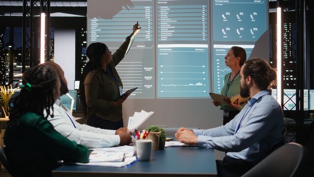 Multiethnic experts team evaluating data performance metrics on projection board, reviewing data insights with investors group in executive session after hours. Corporate event at night. Camera A.