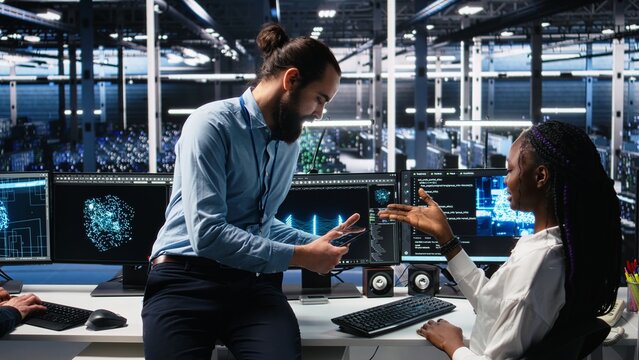 Manager supervising woman in data center coding with generative AI assistance. Supervisor oversees work by african american IT expert leveraging artificial intelligence large language model, camera A