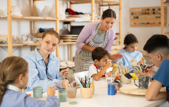 Children paint ceramics with brushes at the table in the creative studio. Teacher shows and helps children at the master class on ceramics