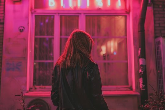 Stylish woman in black leather jacket looking at pink window on urban street, back view at night