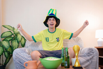 Torcedor brasileiro comemorando em casa enquanto assiste a um campeonato internacional de futebol pela TV. Ele veste a camiseta verde e amarela, vibra no sofá e torce pela vitória do Brasil. © Marcio
