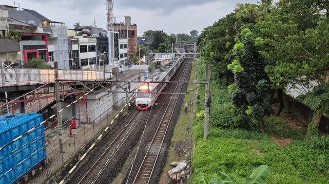 Commuter line train approaching a station in Jakarta, Indonesia. High angle view of urban railway tracks, overhead lines, and city buildings.