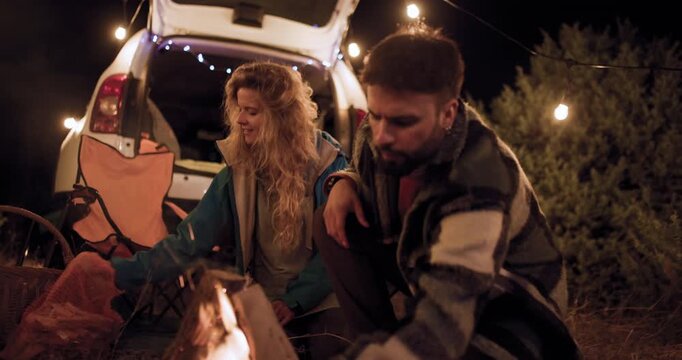 Two friends make a campfire in the woods at night while sharing stories and laughing under the stars with lights in the background