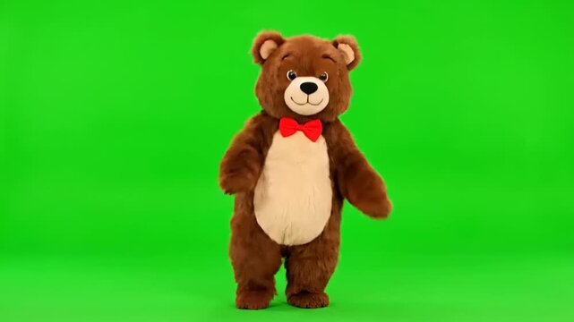 Brown teddy bear mascot dressed in a red bow tie standing and waving on a green screen background, perfect for playful content and children's programming