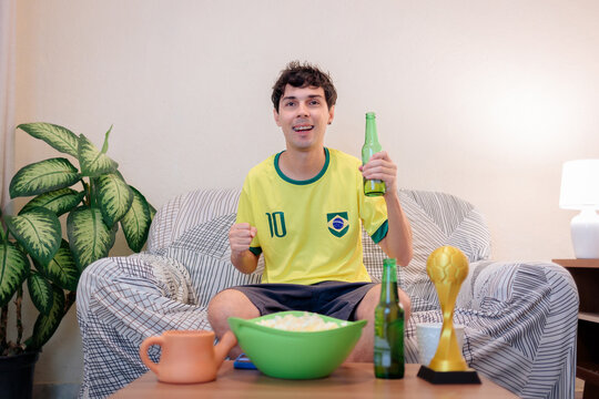 Torcedor brasileiro comemora em casa assistindo jogo internacional na TV. No sof&aacute;, com camisa da sele&ccedil;&atilde;o, ergue cerveja e vibra com pipoca e trof&eacute;u, mostrando paix&atilde;o e alegria.