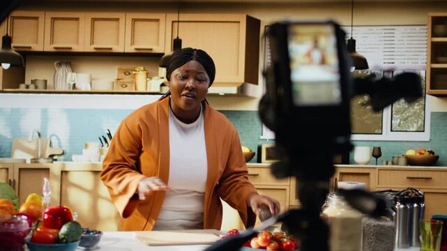 Black woman food vlogger filming culinary content with tripod and camera, behind the scenes of cooking tutorial in home studio kitchen. Influencer ensuring meal prep for online audience.
