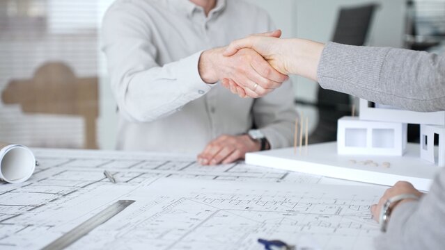 Handshaking between two architects finalizing a construction project deal, business partners agreeing on engineering blueprints and architectural models in an office, close up