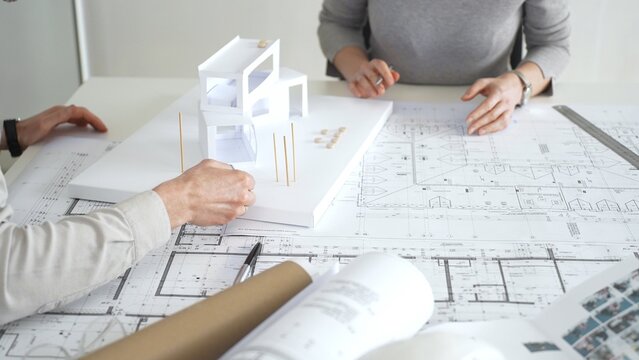 Architects collaborating, examining building blueprints with rulers and pens, developing innovative architectural designs and construction plans around a model house, close up