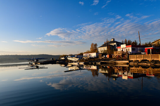 Seaplanes rest at wooden dock beside lakeside buildings, reflected in still waters of Great Slave Lake
