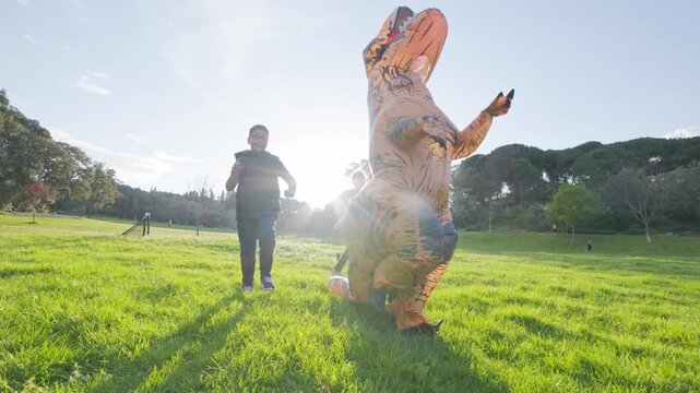 Children chasing and tackling inflatable dinosaur costume in a park