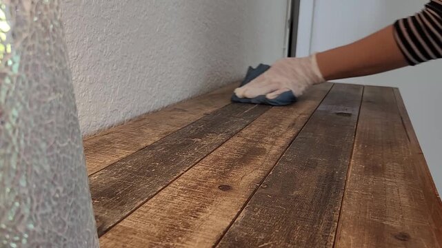A woman wearing gloves wipes a wooden table with a cloth while moving a decorative object. Scene represents cleaning routine, hygiene, and home care.