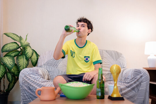 Torcedor brasileiro sentado no sof&aacute; em casa, bebendo uma cerveja enquanto assiste a uma partida internacional de futebol pela TV. Vestindo a camisa da sele&ccedil;&atilde;o brasileira, ele torce pelo Brasil. 