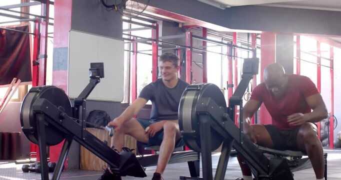Diverse male duo walking into gym, adjusting rowing machine straps, rowing together for cardio