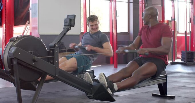 Diverse male friends at gym adjusting straps then rowing together on rowing machines for fitness
