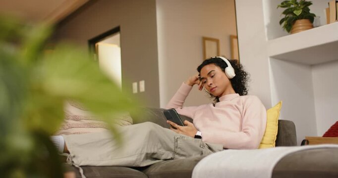 Settling onto sofa, non-binary person scrolling phone to relax, adjusting headphones, gazing upward