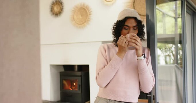 At start non-binary person sipping mug at living room door gazing outside with wood-burning stove