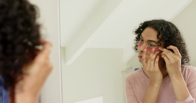 Nonbinary person facing bathroom mirror peeling undereye patches, pressing under eyes for skincare