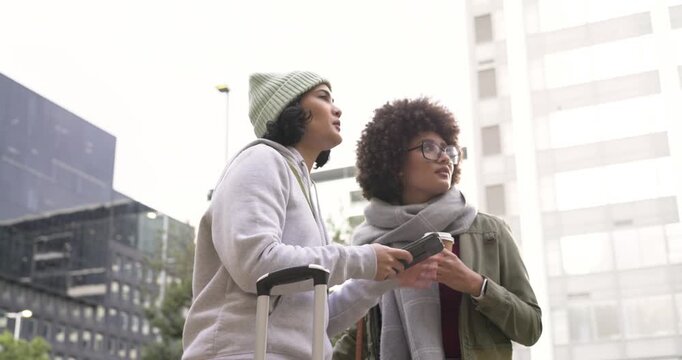 Diverse female friends opening smartphone map on sidewalk with suitcase pointing route and walking