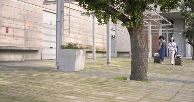 Female friends entering courtyard pulling tan and dark suitcases past tree under canopy, copy space