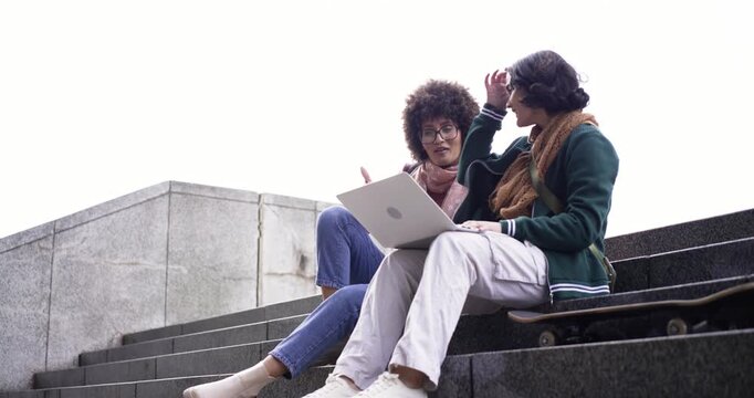 Leaning in to view screen, female friends discussing and typing project on laptop at campus steps