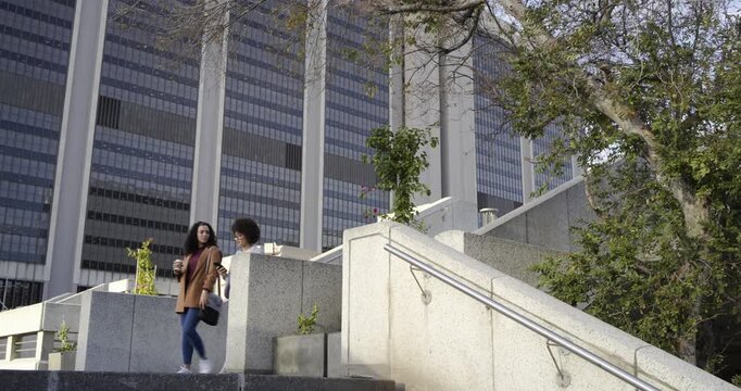 Diverse female friends stepping onto plaza stairs after greeting, sharing phone and sipping coffee