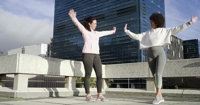 Facing each other on rooftop, diverse female friends warming up with arm sweeps, shoulder pulls