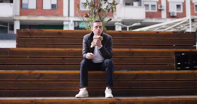 A man sits on wooden steps in an urban park, savoring a tasty snack. The sun shines brightly, and surrounding greenery complements the relaxing atmosphere
