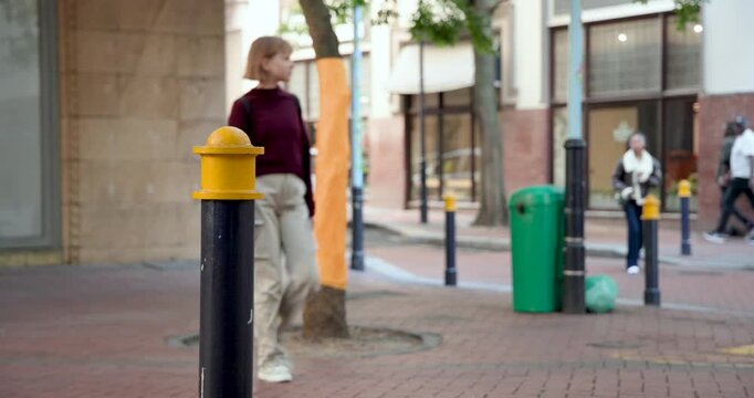 Diverse pedestrians reacting to distant movement walking past black bollard yellow cap on street