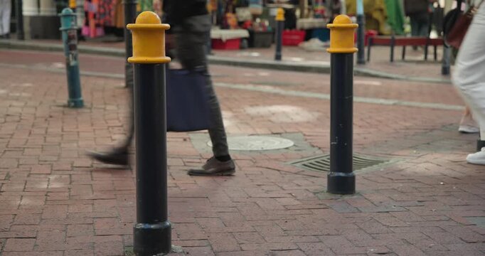 Two short black bollards marking red-brick street while adult shoppers walking past stepping around