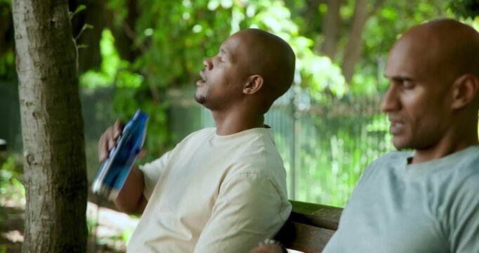 Two African men sitting bench under canopy, catching breath, drinking from bottles, laughing
