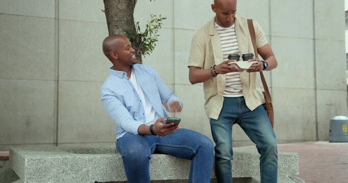 African American men exchanging takeout coffee on arrival, one holding phone on bench by tree