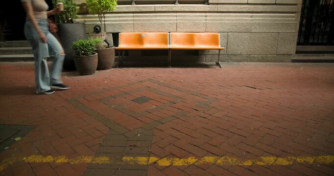 Diverse pedestrians entering frame, walking across brick sidewalk passing planters and orange bench