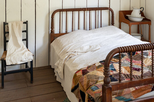 A vintage wooden bed frame, antique wooden chair, nightgown, corner table, jug and basin and bedpan in a small white bedroom. The floors are hardwood. The bedsheets are white cotton and a patch quilt.