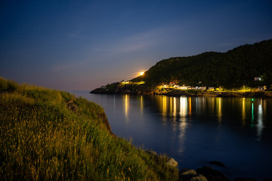 A summer scene of St. John's, Newfoundland, at sunset. The sky is blue with a pale pink hue. The full moon is rising over Fort Amherst lighthouse. The water is reflecting the colourful street lights.