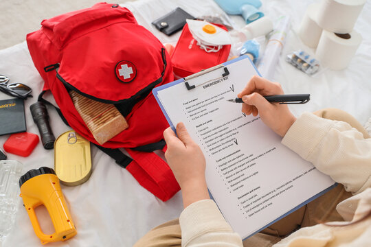 Young woman with checklist packing emergency backpack on bed, top view
