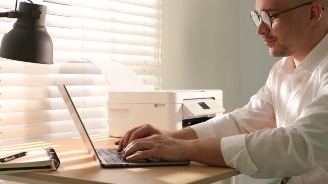 Man using printer while working on laptop at wooden desk indoors