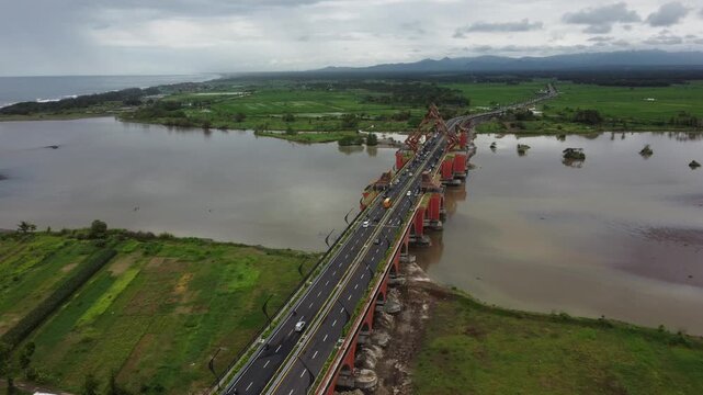 Aerial view of a long bridge crossing a wide river with surrounding green landscape under a cloudy sky.