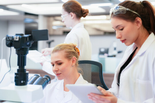 Diverse team of female scientists working in busy digital pathology lab. Researchers analyzing histology slides using microscope, tablet, and visual inspection for medical diagnosis.