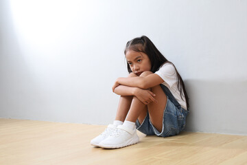 Fototapeta premium Close-up portrait of a young girl sitting on the floor, huddled against a gray wall, feeling deep sadness and helplessness, experiencing bullying, crying.