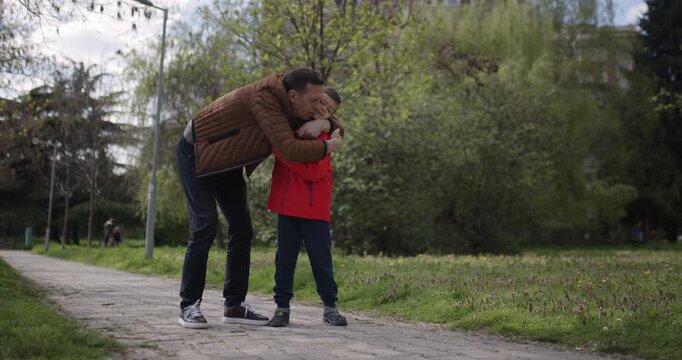 A father and his young son engage in a heartfelt conversation while walking through a peaceful park filled with greenery. The boy listens attentively as they stroll