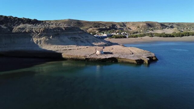 Aerial shot from the transparent green sea towards an abandoned structure in the city of Puerto Piramides Chubut. Rocky reefs and the village in the background are visible.