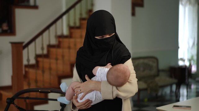 Portrait of a loving mother in traditional muslim clothing, wearing a black niqab and hijab, gently holding her newborn baby in her arms