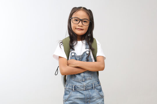 A confident 7-8 year old schoolgirl, folded arms, wearing a backpack, smiling looking at the camera, isolated on a gray background.