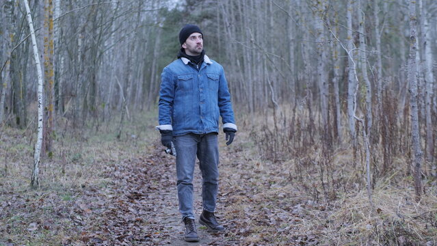 Man in denim jacket and hat pauses on a winter forest trail, holding a metal bottle and looking up at bare birch trees amid dry leaves and quiet solitude