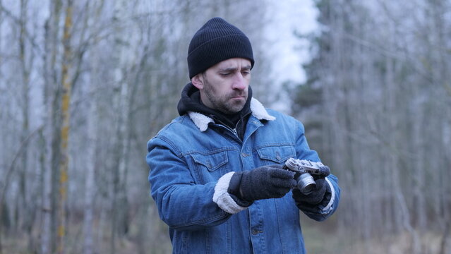 Man in winter clothing wearing a beanie and denim jacket with a sherpa collar, standing in a barren forest and concentrating on adjusting the settings of his vintage rangefinder camera
