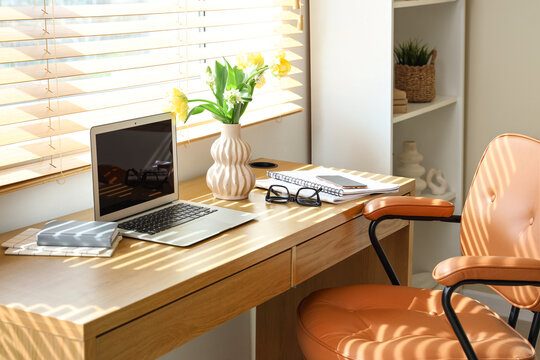Blank laptop and vase with spring flowers on desk near window in office